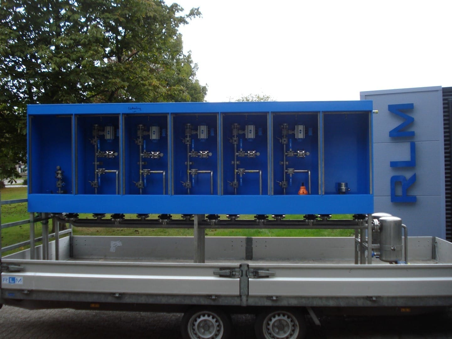 Blauwe doseerkast met vijf vakken, elk met eigen leidingwerk en afsluiters, op een aanhanger voor het pand van RLM