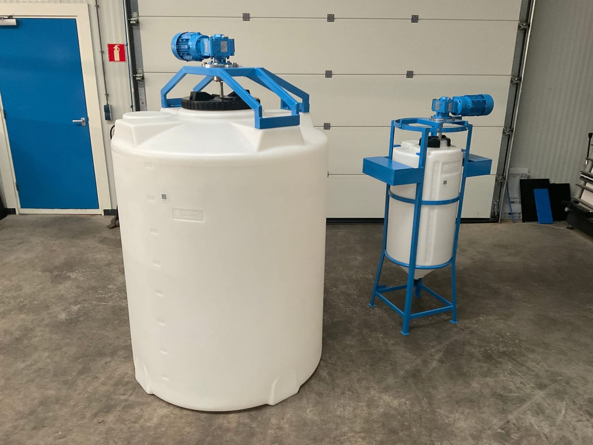 Twee witte ronde tanks met blauwe roerwerken: een grote op de vloer en een kleinere in een blauw frame