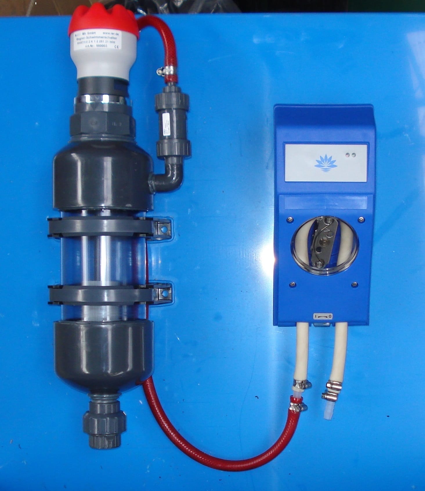 Automatisch hevelvat van grijs PVC met kijkglas en niveausensor met rode kap, naast een blauwe besturingskast met rode slang