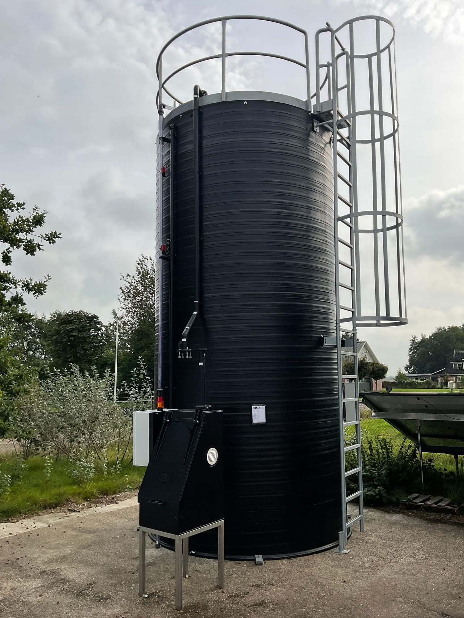 Zwarte staande chemietank buiten opgesteld, met kooiladder, bordeshek en een zwarte doseerkast aan de voet