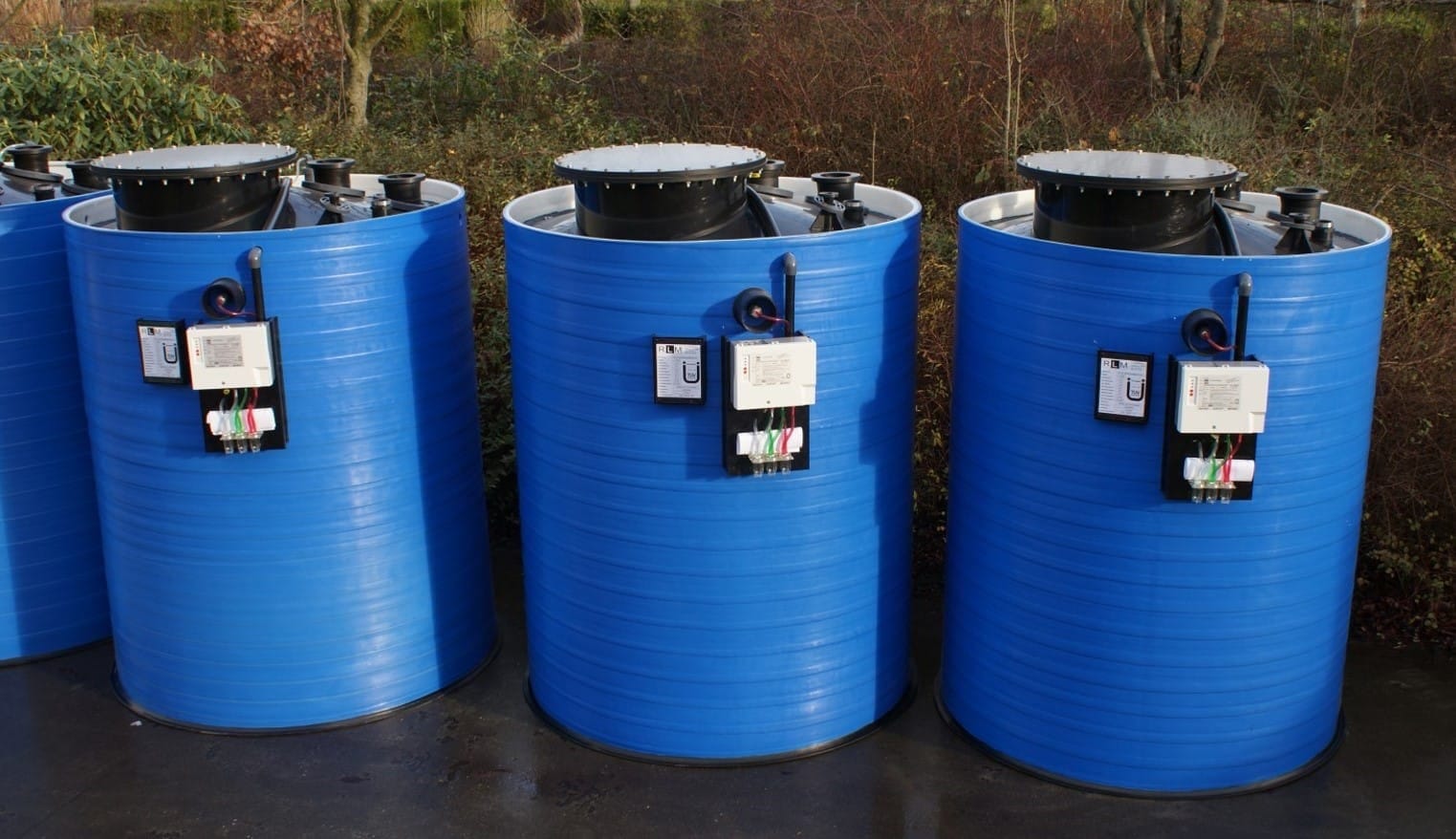Drie blauwe ronde kunststof tanks op een rij buiten, elk met zwart mangatdeksel en een besturingskastje op de wand