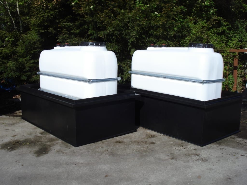 Twee witte rechthoekige PE-tanks met spanbanden, elk in een zwarte PE-opvangbak, buiten voor een coniferenhaag